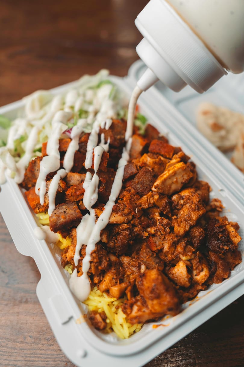 Creamy white sauce drizzled over a takeout container of chicken and lamb over rice with salad.