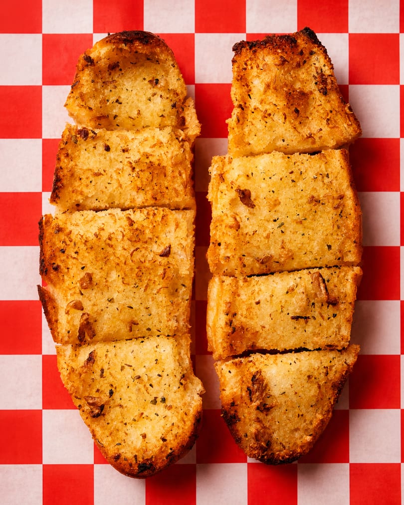 Toasted garlic bread slices with herbs and garlic bits on red and white checkered paper.