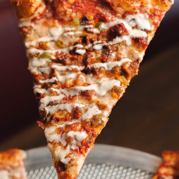 Hand holding up a slice of pizza with meat, green peppers, and white sauce drizzle.