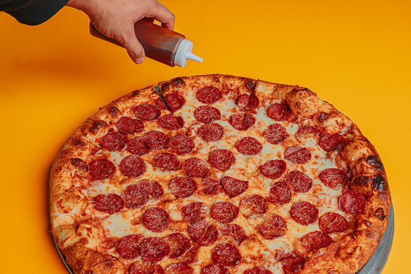 A hand squeezes a sauce bottle over a large, cheesy pepperoni pizza on a yellow background.