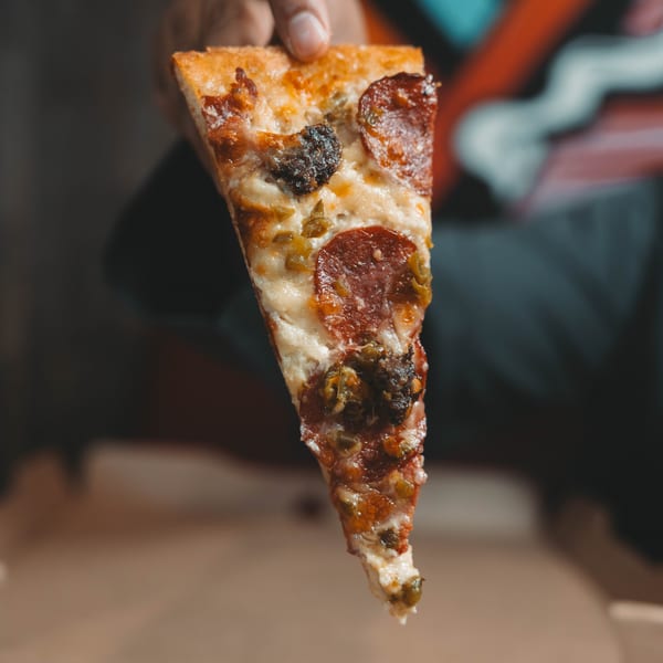 A hand holds up a slice of pepperoni pizza with green peppers and melted cheese.