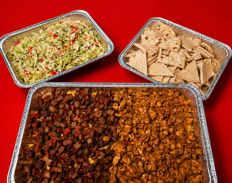 Catering trays filled with diced meat, shredded cabbage salad, and pita bread on a red background.