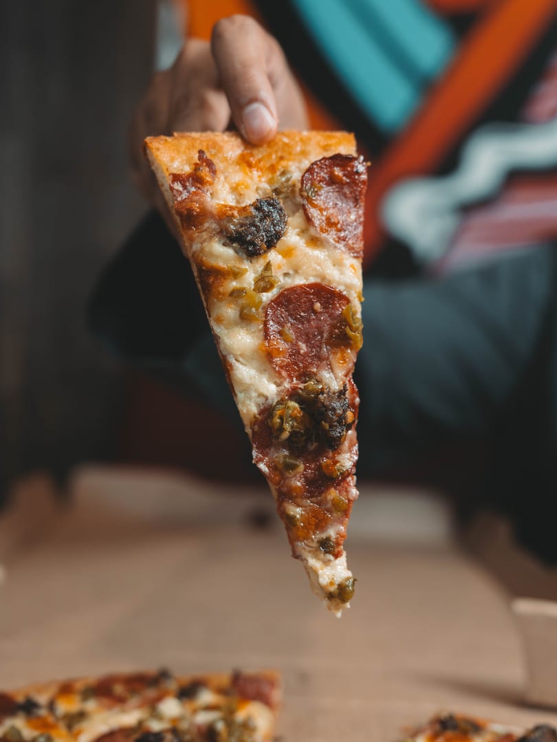 Hand holding a slice of pepperoni pizza with jalapeños and melted cheese.