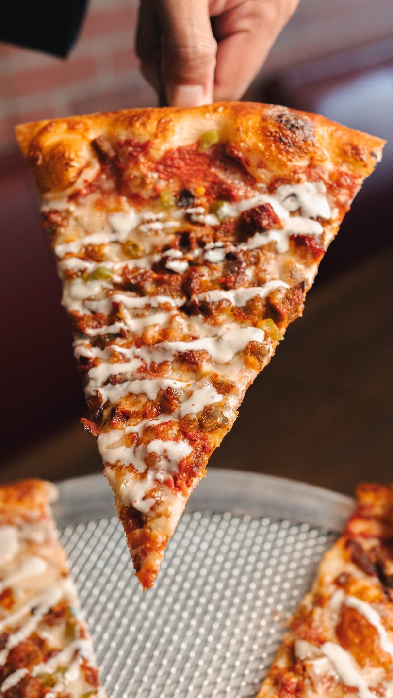 A hand holds up a large pizza slice topped with meat, peppers, cheese, and a creamy white drizzle.